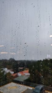 Raindrops on a window with a blurred view of buildings and trees outside, depicting a rainy and overcast day.