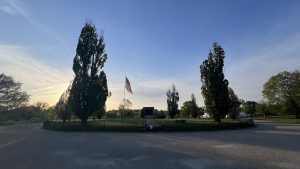帕特森公园（马里兰州巴尔的摩市卡西米尔·普拉斯基将军纪念碑）内，围绕着一个圆形草地公园的黑色铁栅栏，周围长满大树，旁边是石头和金属纪念碑，旗杆上飘扬着美国国旗