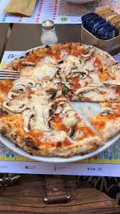 A plate with a partially eaten pizza topped with mushrooms, mozzarella cheese, and tomato sauce is on a table. A salt shaker, packets of sugar, and a fork are visible on the table.