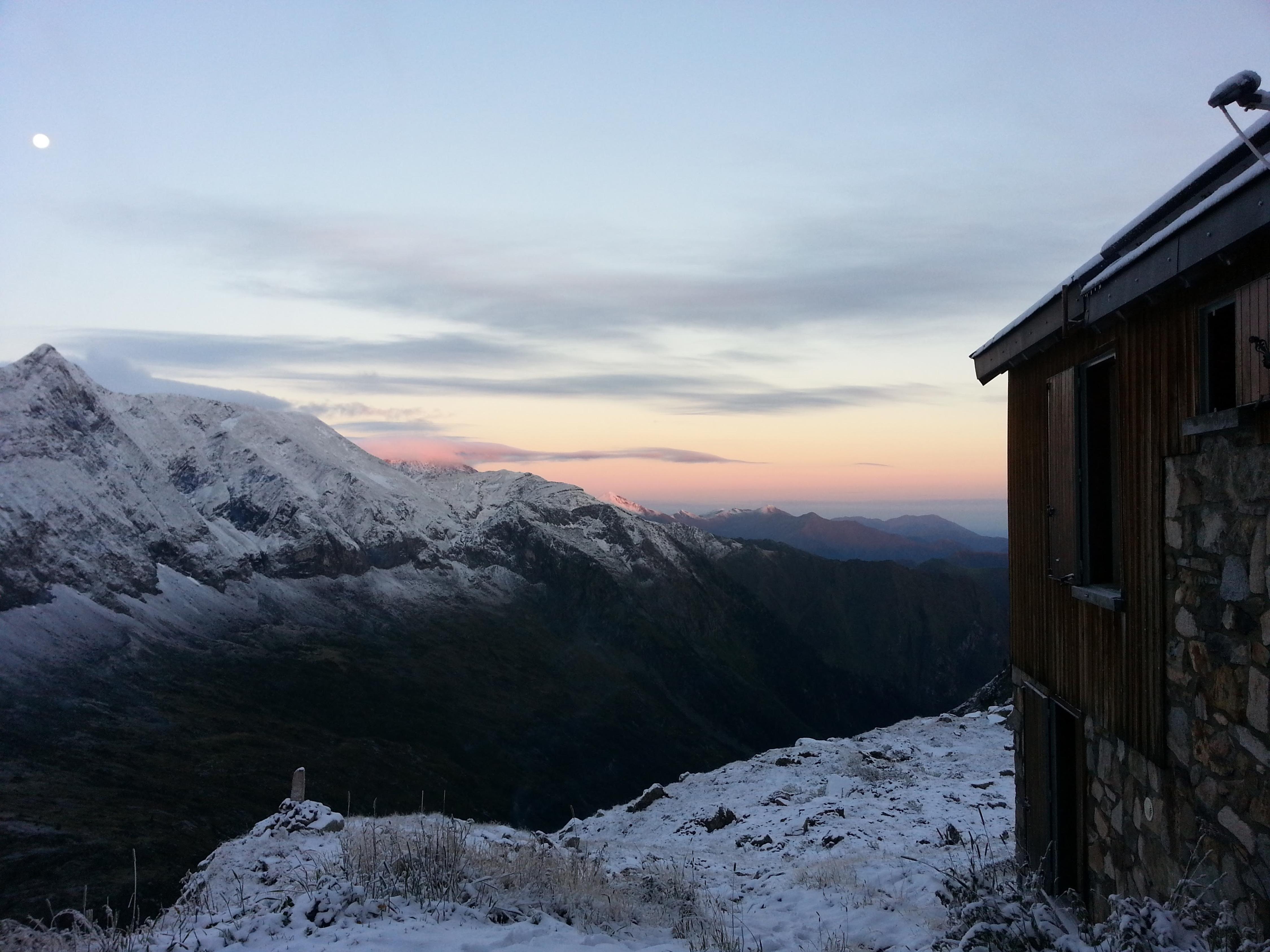 View from Refuge les Estagnous, Mont Valier, at sunrise. Snow-covered mountains against a lightly cloudy blue and pink sky, with a wood and stone building in the foreground.