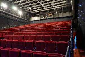 An empty theater with rows of red seats and a partial view of the stage lighting and sound equipment from the back of the room. The walls are dark, with small, blue lights scattered on the sides.