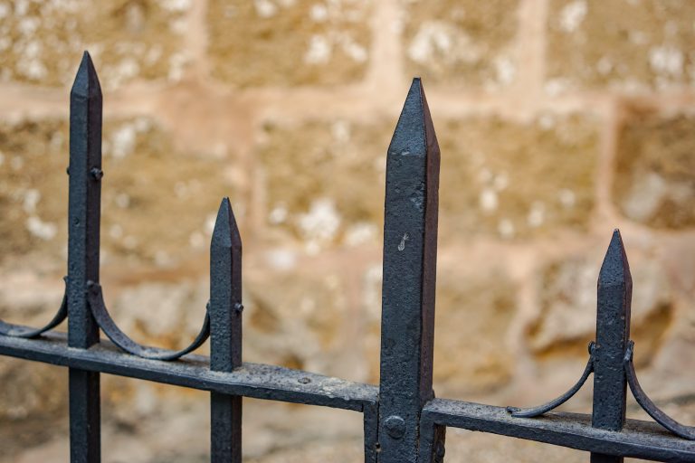 Close-up of a decorative, black, wrought iron fence with pointed tops in front of a blurred, textured stone wall.