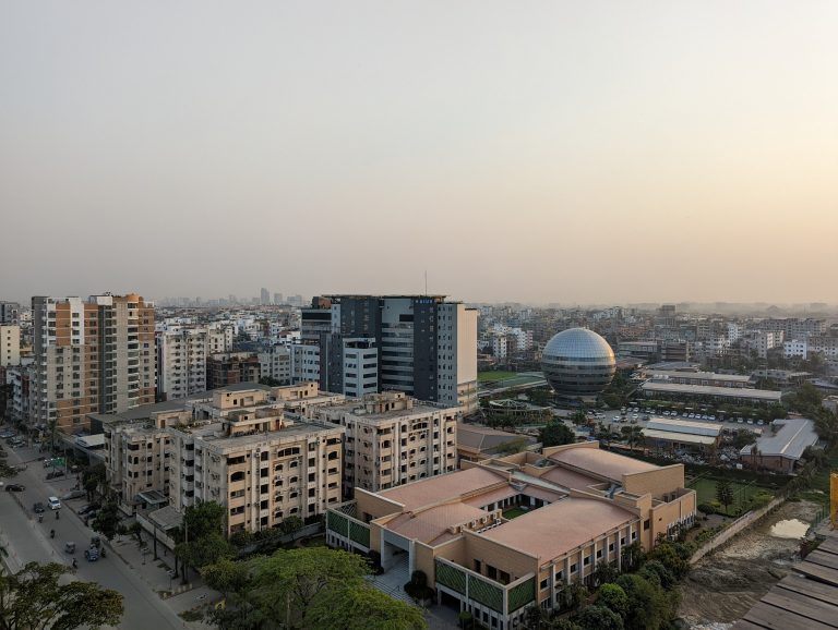 A cityscape with many buildings, including one shaped like a sphere. Cars and pedestrians can bee seen from a distance on the streets.