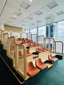  Raised, tiered rows of seats in the Paris airport waiting area. where passengers can sit to watch television. The airport tarmac is visible though the window.
