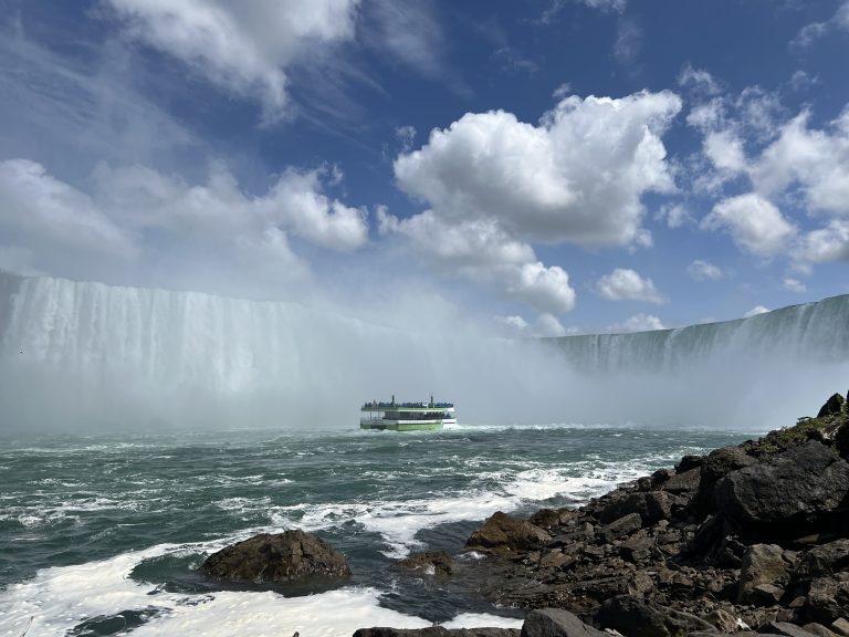 The Maid of the Mist boat at the base of Niagara Falls as seen from the bottom of the falls. Horseshoe falls rises above it. The boat is surrounded by rising mist. Fluffy clouds fill a bright blue sky.