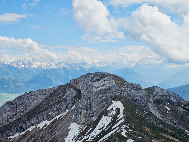 A stunning mountain landscape with rocky peaks partially covered in snow, from Mount Pilatus, Switzerland, under a bright blue sky with scattered white clouds. In the background, additional snow-capped mountains stretch out into the distance, framing a picturesque valley below.