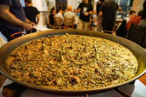 A huge pan of Valencian paella with green beans and chicken drumsticks on a table surrounded by people, some of whom are serving themselves while others are seen chatting and sitting at dining tables in the background.