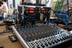 View larger photo: Close-up view of an audio mixing console with various cables and connectors plugged in, placed on a wooden table. In the background, there are additional audio equipment and blurred people.