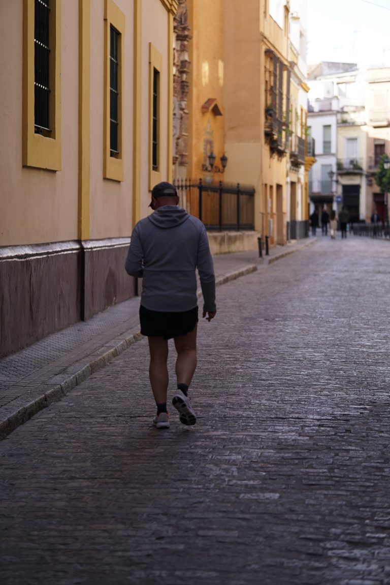 A person wearing a gray hoodie, black shorts, and sneakers walks down a narrow, cobblestone street lined with colorful buildings.