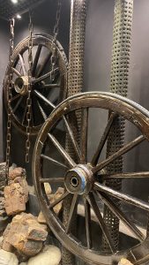 Two large, old wagon wheels hanging vertically by chains in front of perforated metal poles. 