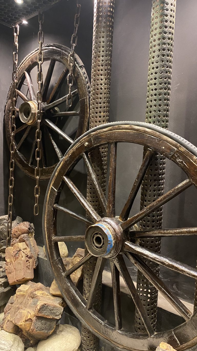 Two large, old wagon wheels hanging vertically by chains in front of perforated metal poles.