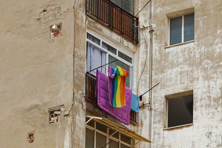 An exterior view of a weathered building wall with visible cracks and patches. A window with a balcony displays a colorful rainbow-striped towel, a purple blanket, and a blue cloth hung out to dry.