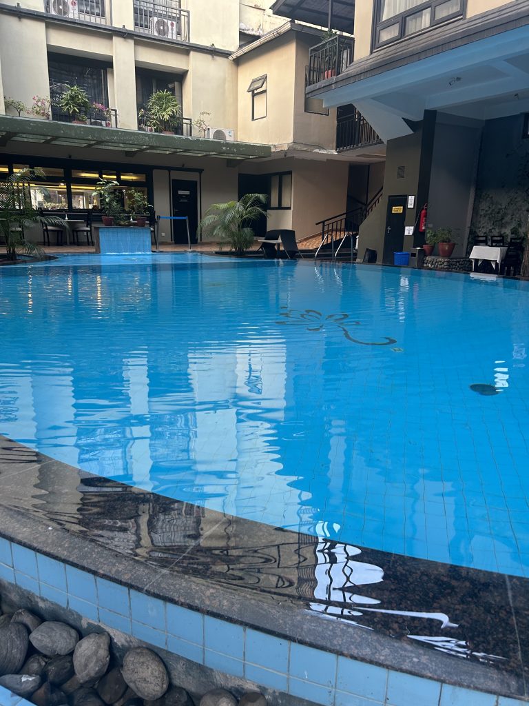 An indoor pool area surrounded by a building with multiple balconies. There are various plants around the pool and seating areas with table and chairs.