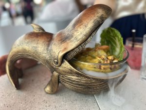 Brass serving dish in the shape of a whale, mouth open with a bowl of ceviche, and dry ice vapor spilling out of the whale's mouth