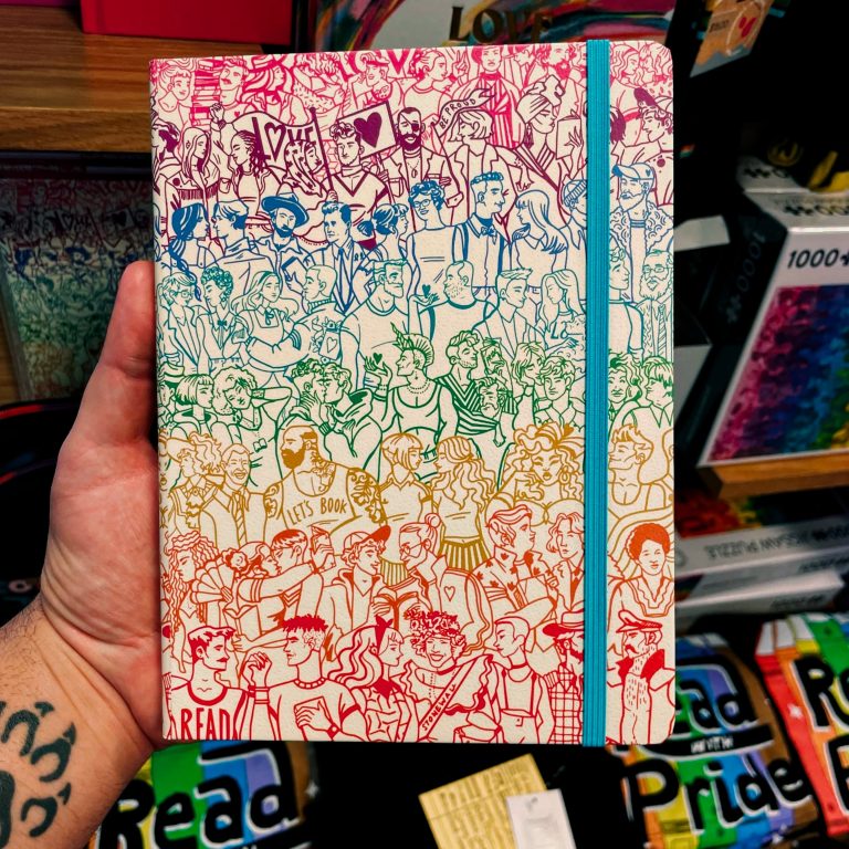 A hand with a wolf paw tattoo holding up a notebook in front of a store shelf. The shelf has shirts that say “Read With Pride” on them. The notebook has drawings of a variety of human body types, lined in colores that make a rainbow gradient