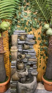 A decorative water fountain featuring stone-like structures and ceramic jugs with cascading water. The background is adorned with green leafy vines and flanked by large artificial palm trees in terracotta pots.