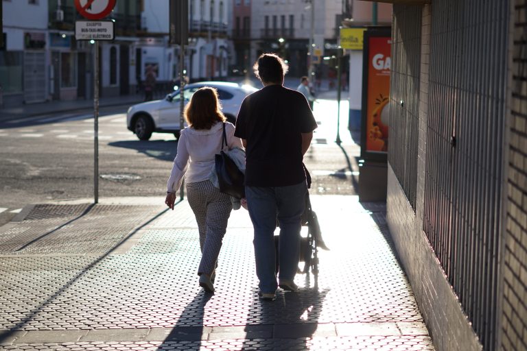 A couple walks sunlit city sidewalk pushing a stroller, with their backs to the camera. The street is lined with buildings, and a car is visible in the background. The sun casts long shadows and a colorful glow on the pavement.