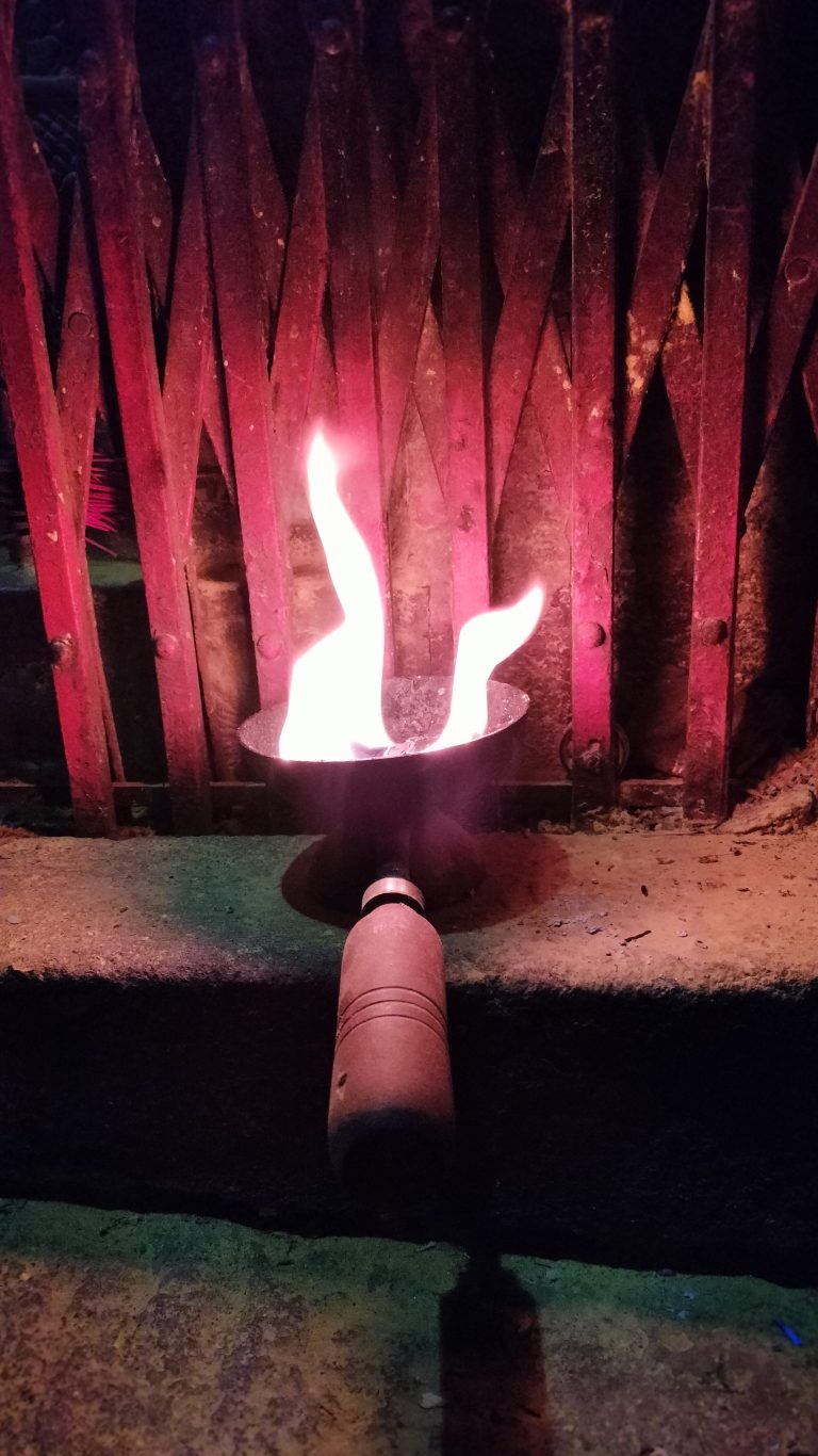 A traditional metal oil lamp with two burning flames positioned in front of a rustic metal gate or barrier, emitting a warm glow. The lamp has a wooden handle extending towards the bottom of the image.