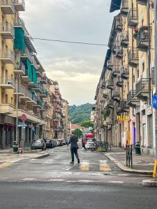 View larger photo: A man walks down a street in Torino, Italy. Buildings rise up either side of the street. Cars are parked on either side. The mountains can be seen in the distance.