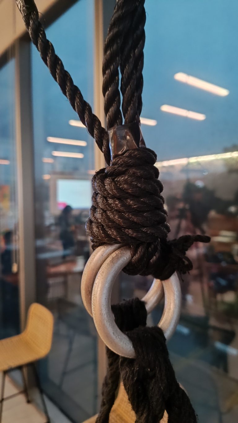 A close-up photo of a thick black rope tied around metal rings, secured with a metal clamp. The background features a blurred indoor space with chairs, large windows, and reflected lights.