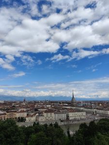 The photo depicts a cityscape under a partly cloudy sky. The city in view is torino, italy. The prominent structure with a tall spire in the center of the image is the mole antonelliana, an iconic building in turin. The city is characterized by a dense arrangement of historic buildings with red-tiled roofs and orderly streets.