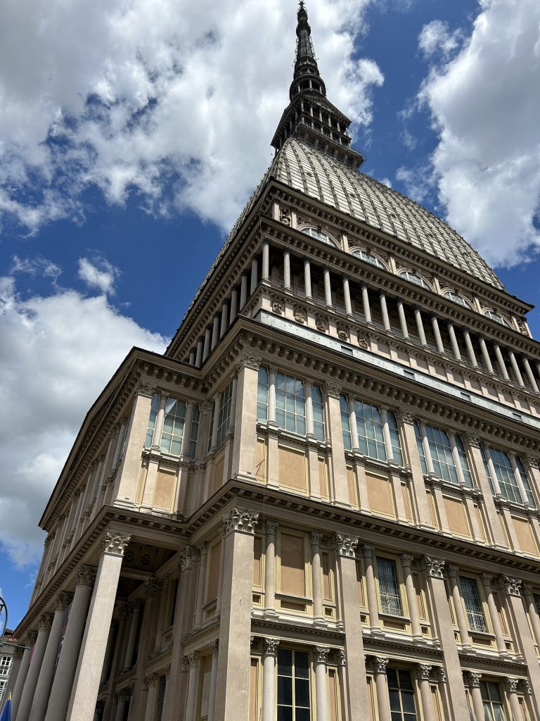 A photo of the Mole Antonelliana building in Torino taken from below, showing the top of the building with a sunny sky as the background.