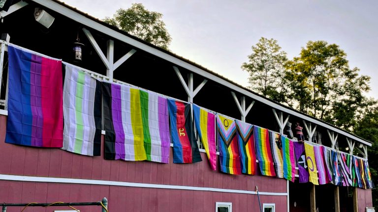 A series of various pride flags for different communities hung up along the second floor of a red barn building.