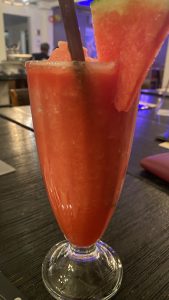 A tall glass of watermelon juice, garnished with a watermelon slice and a brown straw. The drink is set on a dark wooden table.