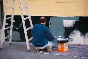 A person is painting the lower part of an exterior wall with a brush. The wall has a yellow upper section and a black lower section, with some areas of the black section being repainted. 