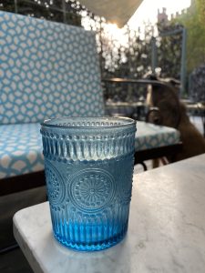 A blue textured glass sits on a white marble table, with a patterned blue cushion chair and a sunlit garden in the background.