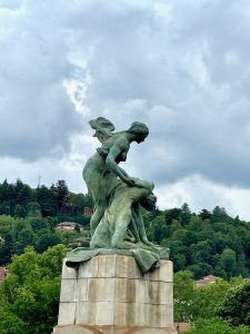  A bronze statue depicting a dynamic scene of two intertwined figures, one appearing to support or rescue the other, mounted on a stone pedestal. This statue is located on Umberto Bridge in Torino, Italy. The background features lush green trees and hillside houses under a cloudy sky.
