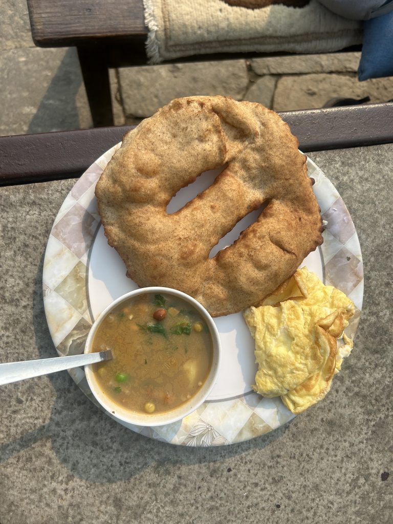 A plate containing a large piece of gurung bread, a folded omelet, and a bowl of soup with visible vegetables and herbs.