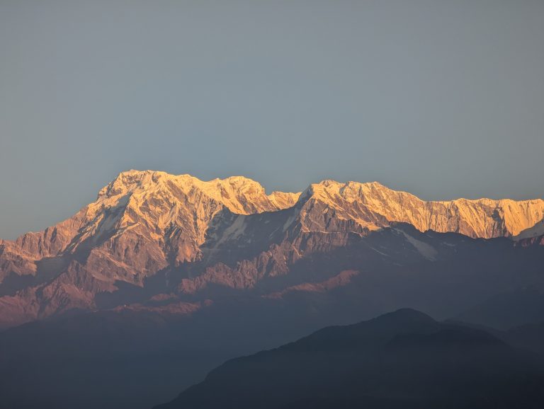 Snow-covered mountain peaks bathed in a warm golden hue, towering against a clear, pale sky. The lower slopes are rugged and shaded, creating a dramatic contrast between light and shadow.