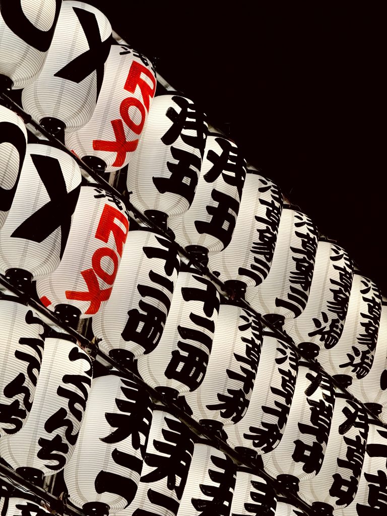 A close-up view of traditional Japanese lanterns, arranged in neat rows against a dark background. The lanterns are white with bold black and red characters, creating a striking contrast. The repetitive patterns and cultural symbols add an intriguing visual appeal.