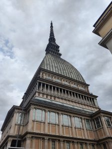 

A photograph of the Mole Antonelliana, a tall and iconic building in Turin, Italy, featuring a distinctive spire and elaborate architectural details, set against a cloudy sky.