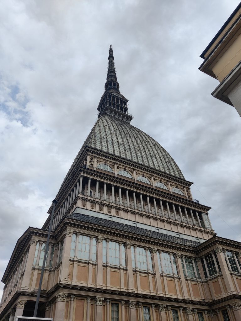 A photograph of the Mole Antonelliana, a tall and iconic building in Turin, Italy, featuring a distinctive spire and elaborate architectural details, set against a cloudy sky.