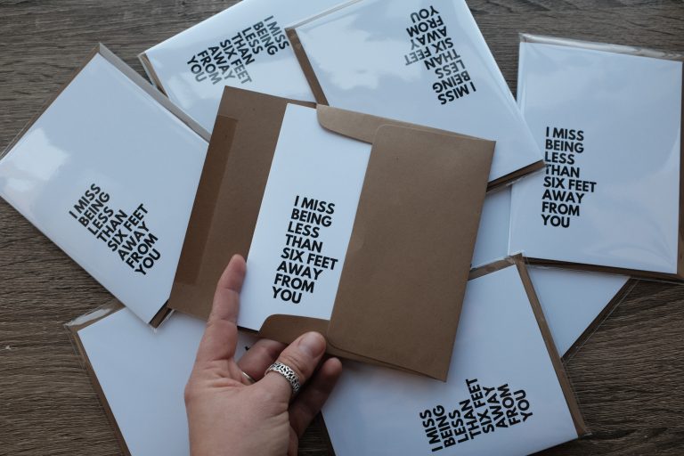 The same white card scattered around with the phrase in all caps in black writing “I miss being less than six feet away from you”. There are eight cards in view with one in the envelope with a hand holding it open.