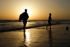 Silhouettes of two people on a beach at sunset, with one person standing with a towel wrapped around them and the other walking towards a ball near the water's edge. The sun is low on the horizon, casting a golden glow on the wet sand and waves.