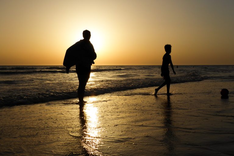 Silhouettes of two people on a beach at sunset, with one person standing with a towel wrapped around them and the other walking towards a ball near the water’s edge. The sun is low on the horizon, casting a golden glow on the wet sand and waves.