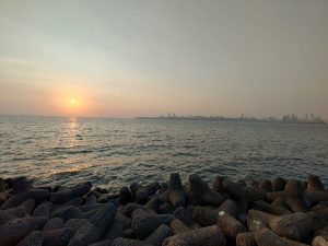 View larger photo: A serene sunset view at Juhu Beach in Mumbai, India. The sun is low on the horizon, casting a warm glow over the ocean. The silhouette of Mumbai's skyline is visible in the distance, while the foreground features a rocky shoreline with large, dark tetrapod structures.