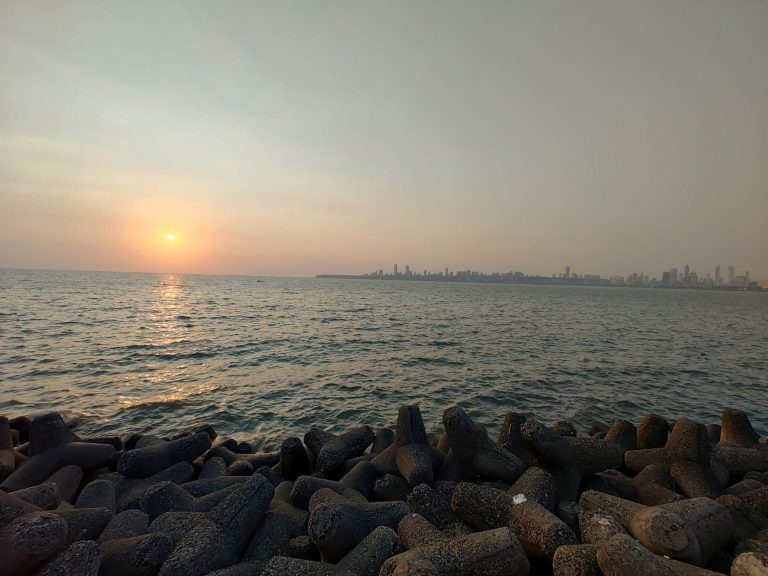 A serene sunset view at Juhu Beach in Mumbai, India. The sun is low on the horizon, casting a warm glow over the ocean. The silhouette of Mumbai’s skyline is visible in the distance, while the foreground features a rocky shoreline with large, dark tetrapod structures.