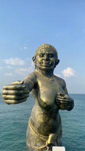 A large bronze statue of a mythological figure extends an arm outward, standing against a backdrop of calm sea and a clear blue sky with scattered clouds.