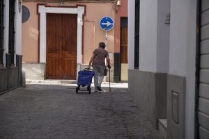 An elderly person using a cane and pulling a blue cart walks down a narrow cobblestone street lined with buildings. In the background, there are wooden doors and a one-way traffic sign on a wall.