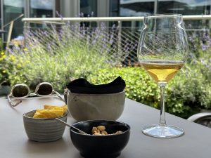 View larger photo: A patio table holds a glass of Aperol Spritz, corn chips, mixed nuts with a spoon, black napkins in a ceramic container, and sunglasses, with a lush garden and blooming lavender in the background.