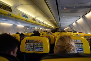 View larger photo: View of an airplane cabin from the back, showing rows of yellow seats occupied by passengers with safety instructions and menu details displayed on the seat backs.