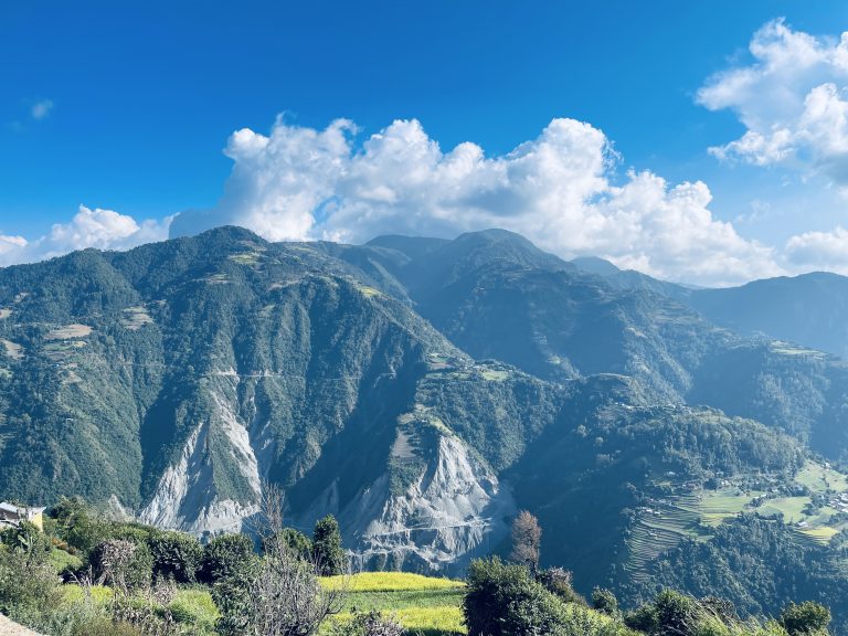 Long view across a valley to tree covered mountains on the other side with fluffy white clouds in a blue sky above.