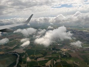 A patchwork of farms and grass as seen from a plane. The wing of the plane is to the left of the frame, and fluffy clouds hover above.