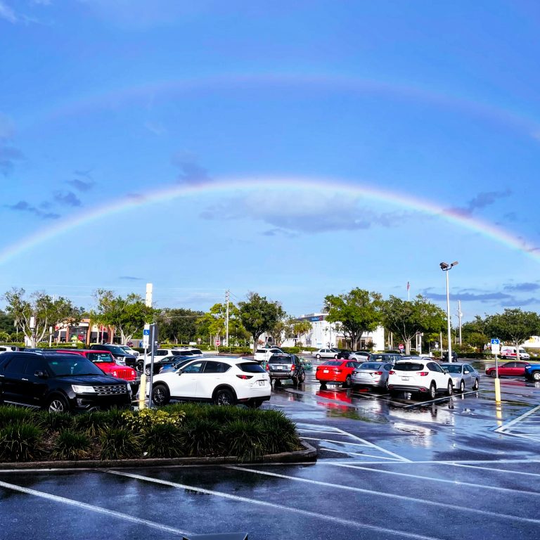 A double rainbow over a blue sky as seen from a parking lot. The asphalt is wet and reflective.