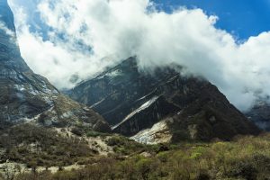 一种山地景观，陡峭的岩石斜坡部分被雪覆盖。山顶被翻腾的白云遮住了。前景是灌木丛和光秃秃的灌木丛的混合。上面的天空大部分是蓝色的，有一些零星的云