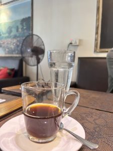 View larger photo: A glass of black coffee on a saucer, with a spoon next to it and a glass of water beside it.